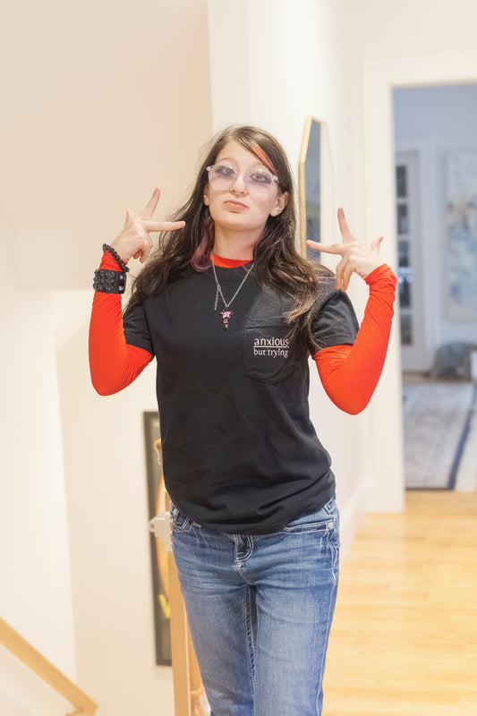 A teen girl posing indoors wearing a black graphic anxious but trying pocket tee t-shirt with red long-sleeve undersleeves, blue jeans, and clear-framed glasses. She is making a peace sign with both hands.