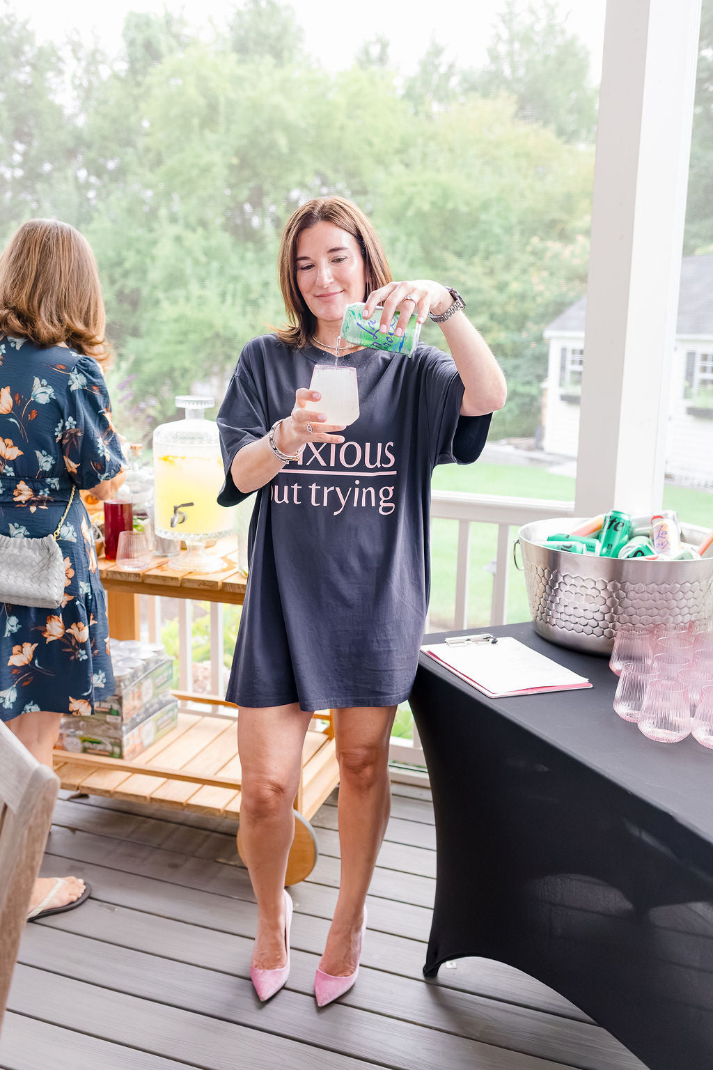 A woman on a porch wearing a grey oversized graphic T-shirt with the white text 'anxiou but trying' and pink heels, pouring a drink from a green bottle into a glass. Another woman in a floral dress stands nearby, and tables with drinks and a basket are visible in the background.